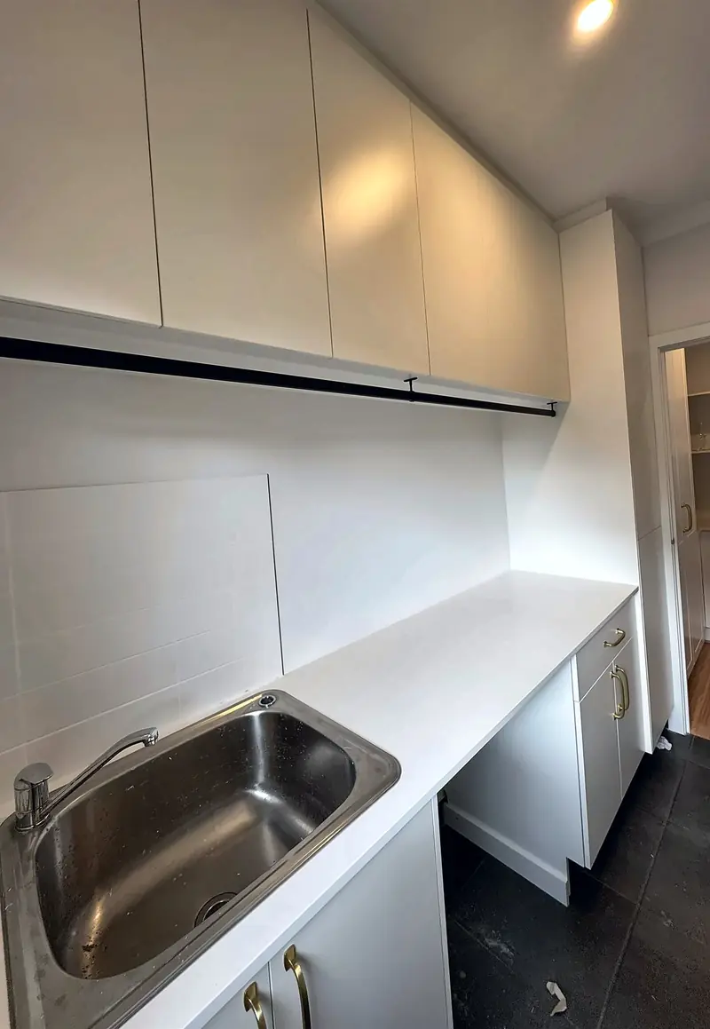 White laundry with a stone bench, tub, overhead cupboards and a hanging rail, by Maya Cabinets in Melbourne